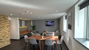 Ein moderner Besprechungsraum mit Holztisch, grauen Stühlen, einer Ziegelwand als Blickfang, einer Couch und einem Fernseher, der einen Comedy-Sender zeigt.