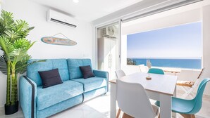 Un salón moderno con un sofá azul, una mesa de comedor con sillas, una planta en maceta y vista al mar a través de un ventanal.