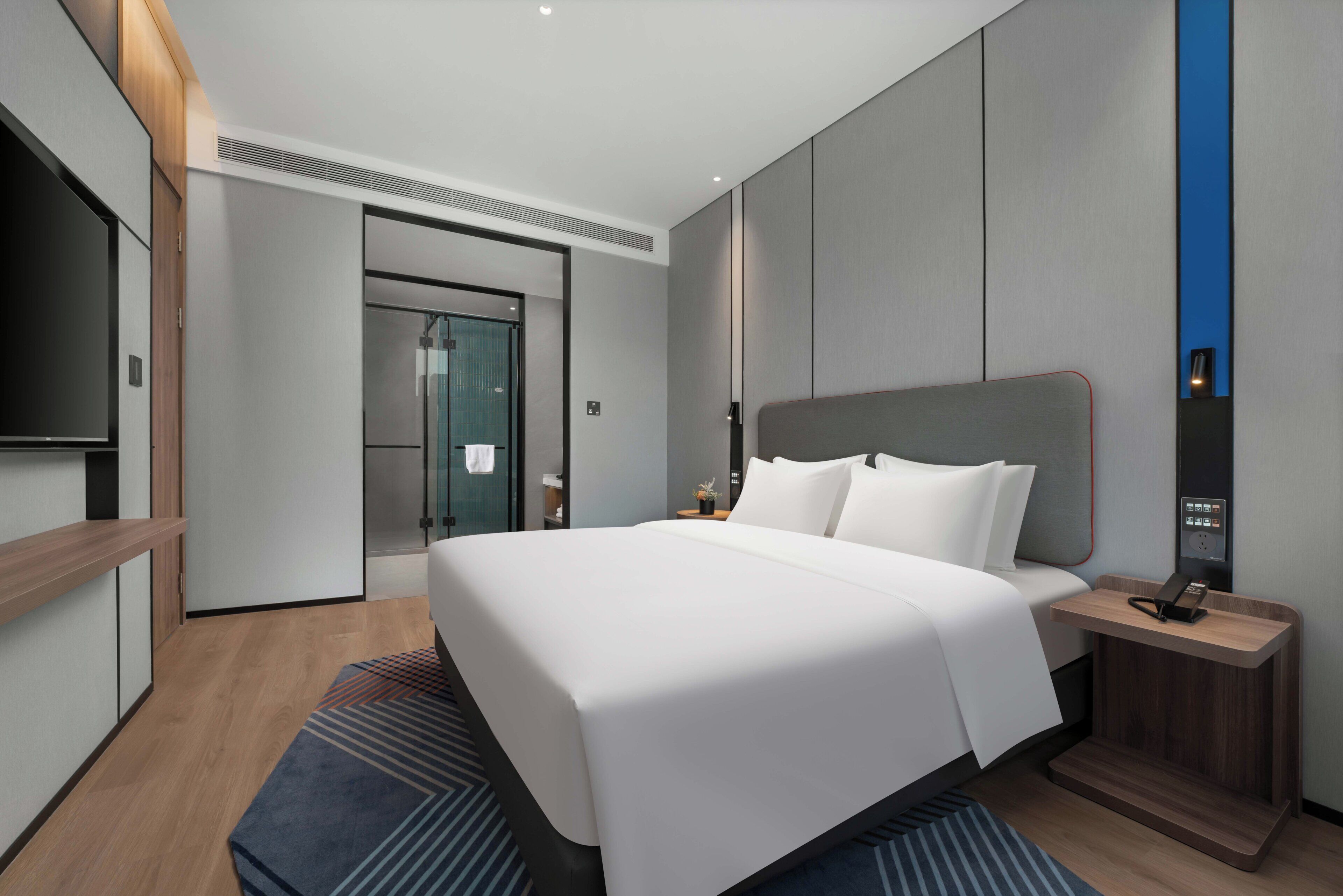 Une chambre d’hôtel moderne dotée d’un grand lit, d’une table de chevet avec un téléphone, d’un téléviseur intégré et d’une porte en verre donnant sur la salle de bain.