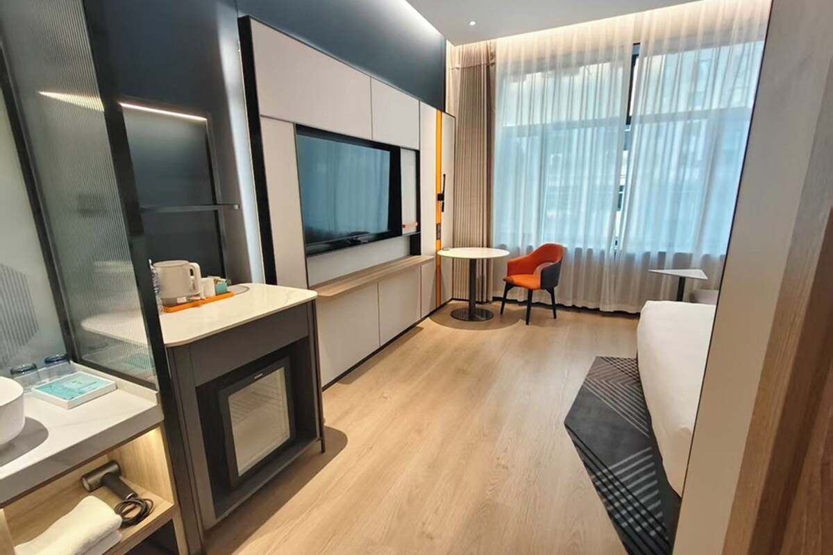 A modern hotel room with a kitchenette, a flat-screen TV, a round dining table, and a comfortable seating area with a view of the city.