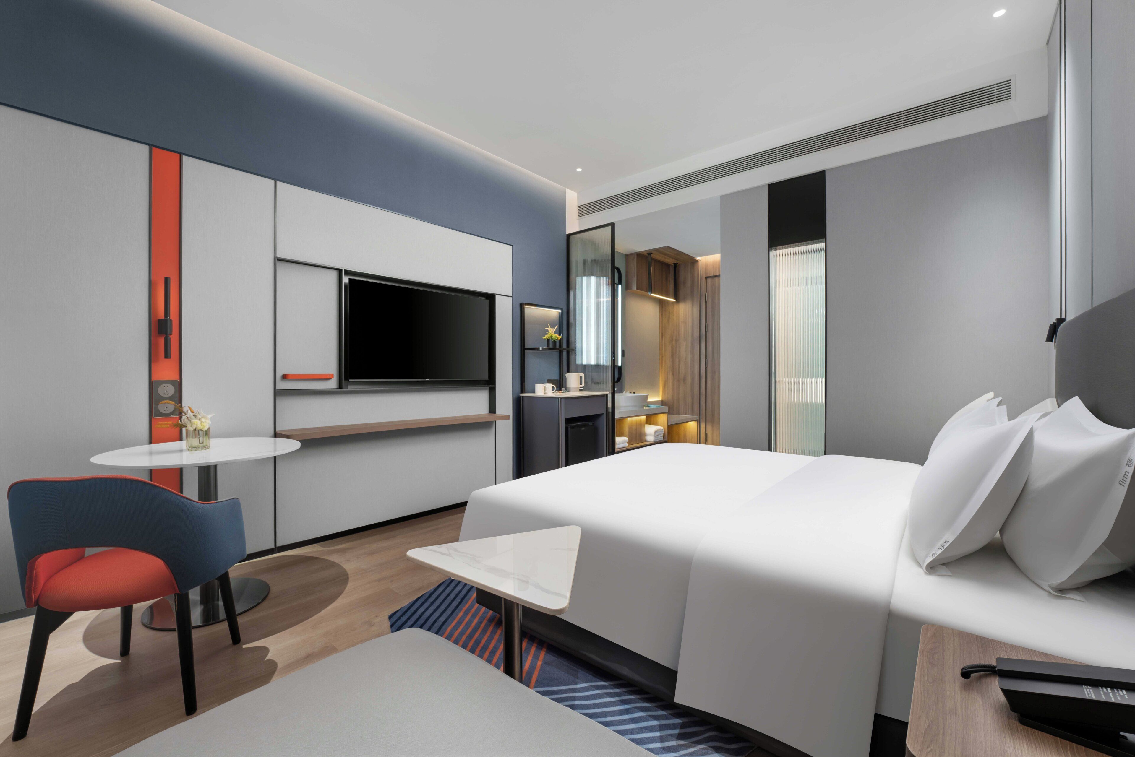 Una habitación de hotel moderna con una cama, un pequeño escritorio, una silla, un televisor y un minibar.