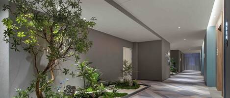 A modern hotel corridor with a central garden, neutral walls, and patterned carpet.