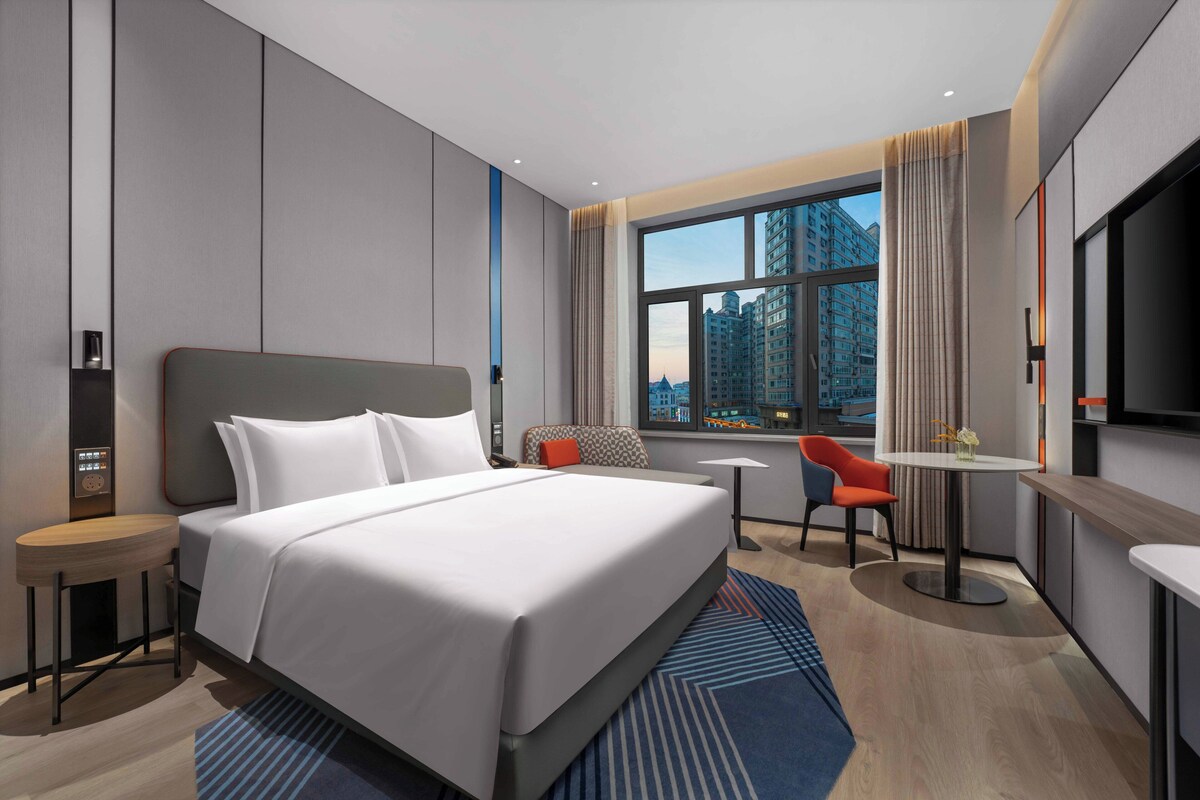 A modern hotel room with a large bed, a desk, a chair, a small table, and a TV.