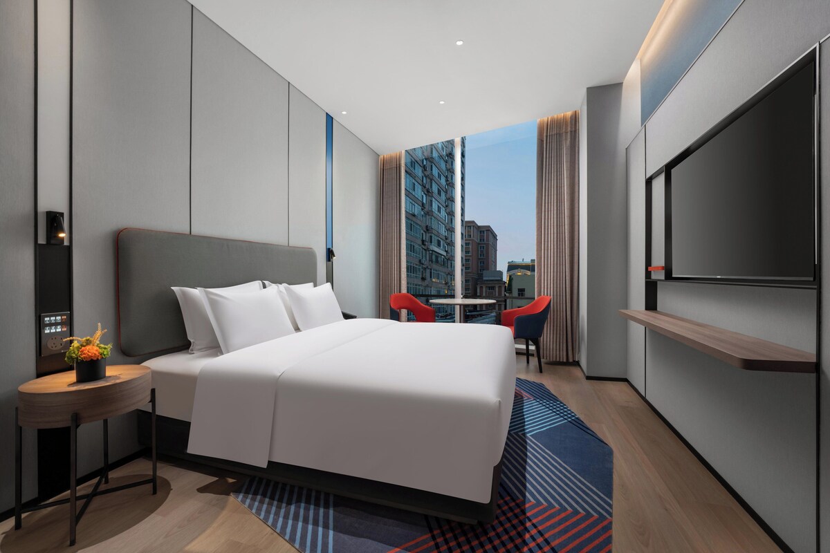 A modern hotel room with a large bed, a TV, a small table with a plant, and a view of the city through large windows.