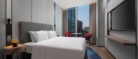 Una habitación de hotel moderna con una cama grande, televisión, una mesita con una planta y vistas a la ciudad a través de amplios ventanales.