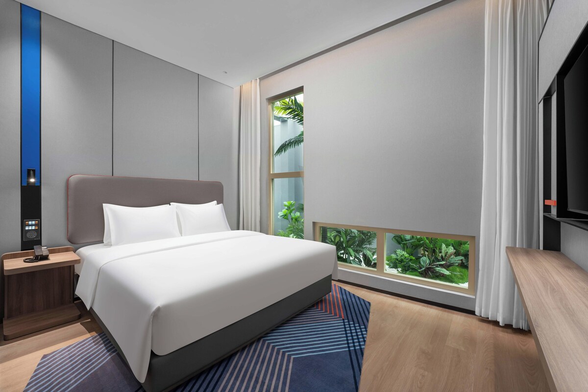 Quarto de hotel moderno com uma cama grande, mesa de cabeceira, TV e janela com vista para vegetação.