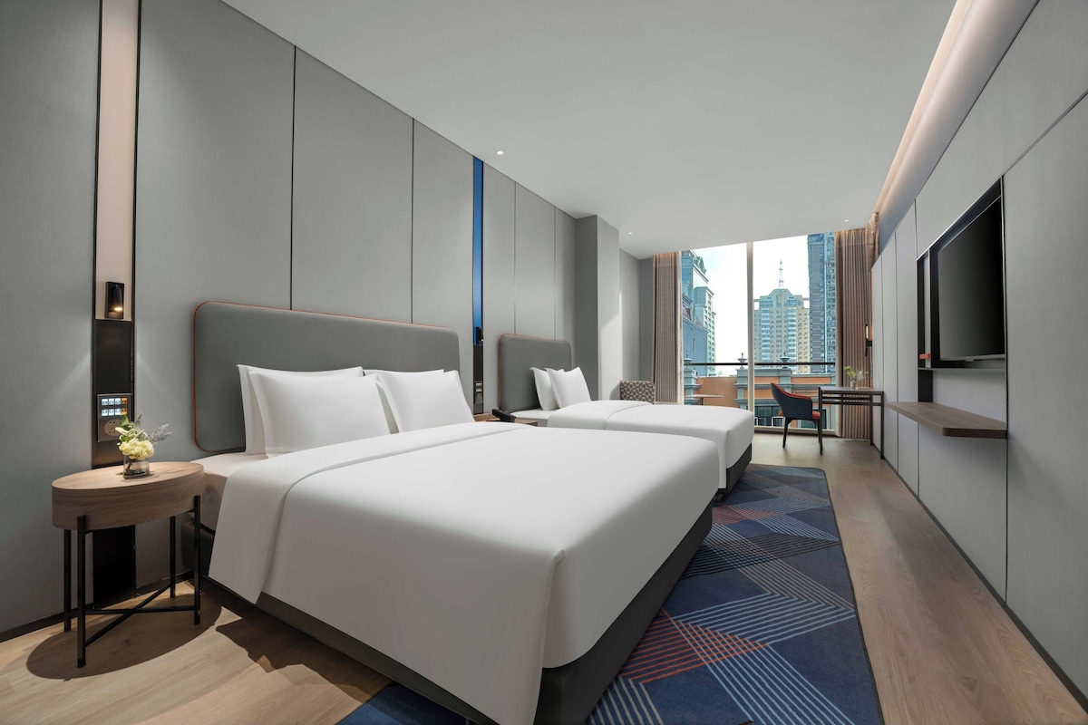 A modern hotel room with two beds, a small table with a vase, a TV, and a city view.