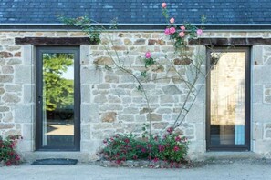 Traditional stone building with a symmetrical facade, featuring two doors and windows, set amidst a garden with flowers and climbing plants.