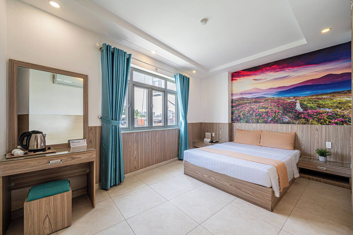 A modern hotel room with a wooden desk, a bed, a large window with curtains, and a vibrant landscape painting on the wall.