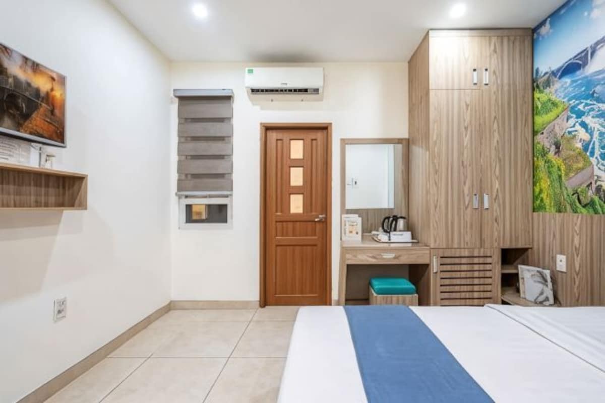 A bedroom with a bed, a desk, a wardrobe, an air conditioning unit, and a painting on the wall.
