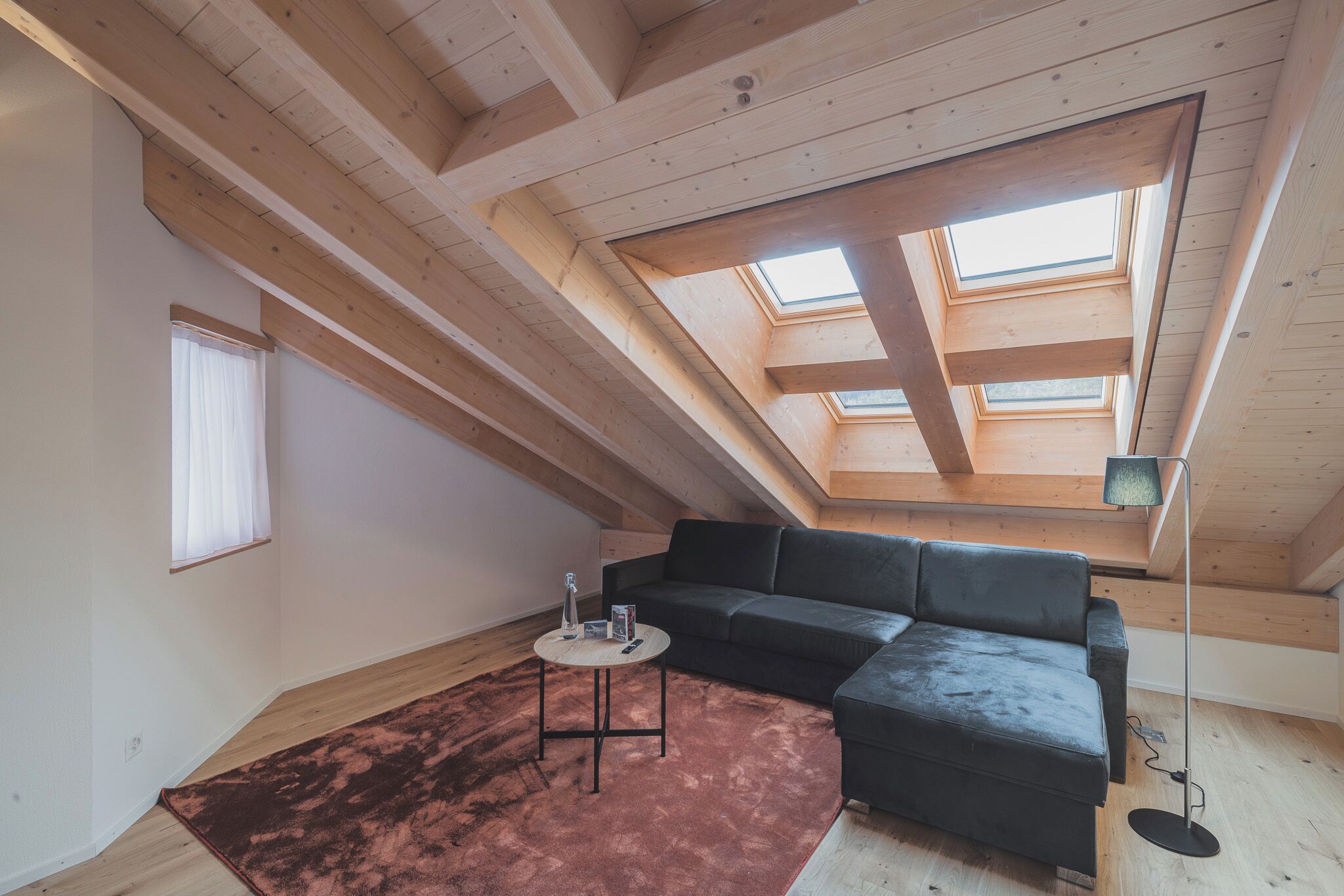 A modern loft with exposed wooden beams, a sectional sofa, a round coffee table, and skylights.