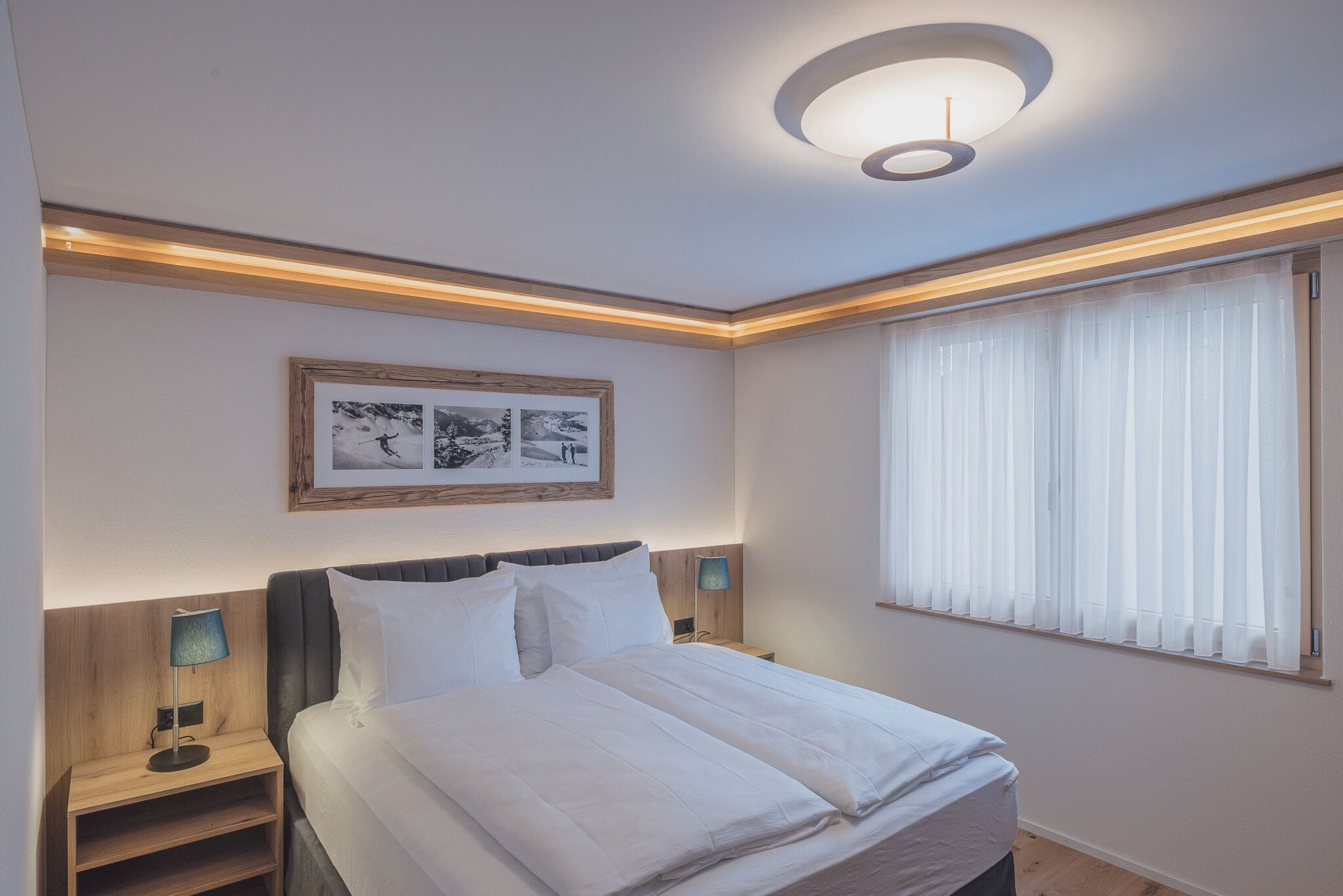 Een keurig opgemaakt slaapkamer met een bed, nachtkastjes, een lijst met een afbeelding aan de muur en een raam met gordijnen.