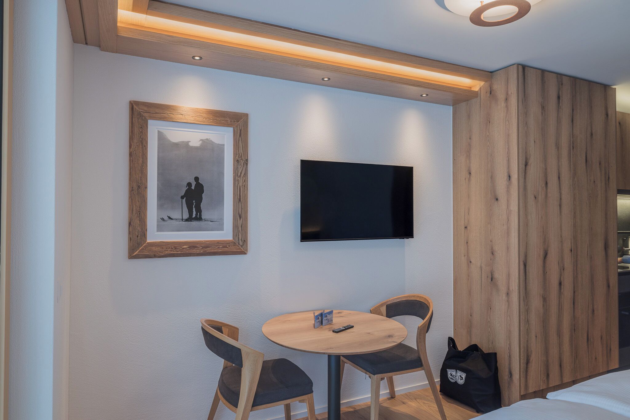 Ein Hotelzimmer mit einer holzverkleideten Wand, einem an der Wand montierten Fernseher, einem gerahmten Bild, einem runden Holztisch mit zwei Stühlen und einer schwarzen Tasche am Boden.