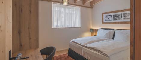 A bedroom with a bed, a desk, a chair, a window with sheer curtains, and a framed picture on the wall.