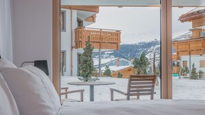 A hotel room with a bed, a round table, and two chairs. The room has large windows offering a view of a snowy mountain landscape. - Val Saraina Resort Apartments Lenzerheide (Lantsch-Lenz)