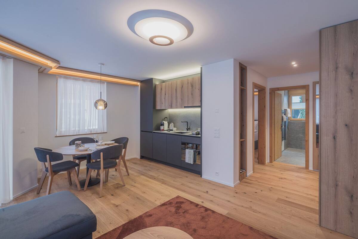 A modern kitchen with wooden cabinets, a dining area with a table and chairs, and a ceiling light fixture.
