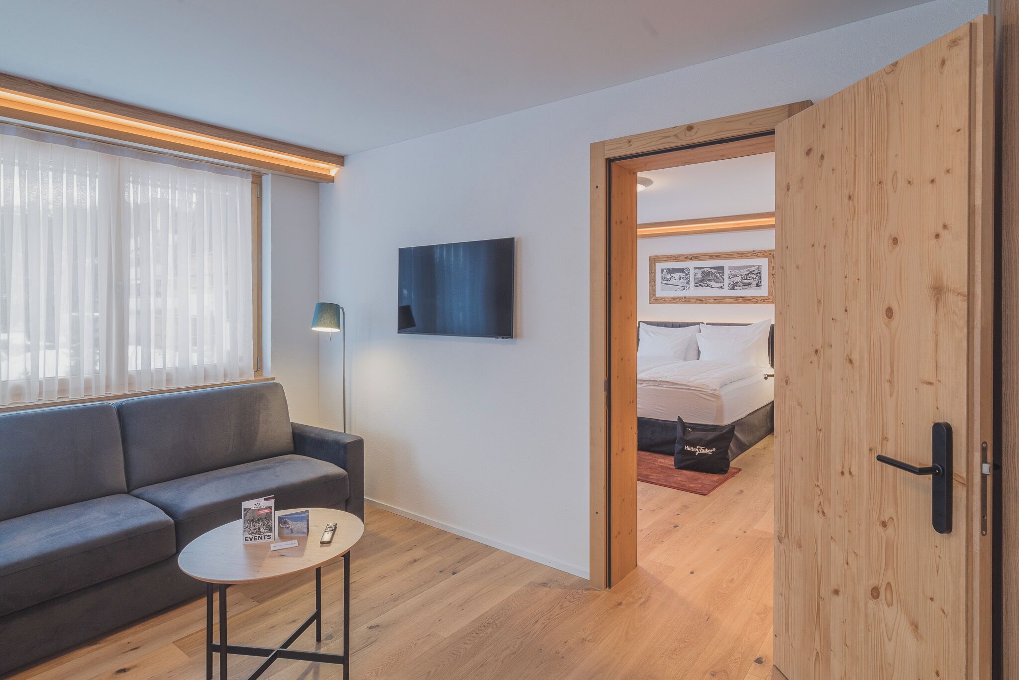 Une pièce moderne avec un canapé gris, une petite table ronde sur laquelle reposent des magazines et une télécommande, un téléviseur fixé au mur, et une porte en bois donnant sur une chambre à coucher équipée d’un lit et d’une table de chevet.