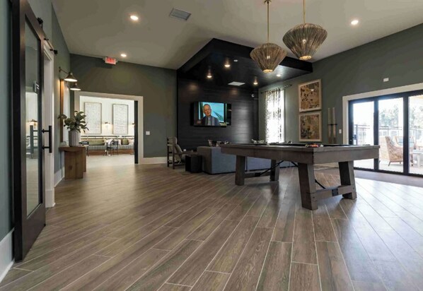 A spacious living area with a pool table, a television, and a large window offering a view of the outdoors. - Landing | Stylish 2BD, Clubhouse, Gym (Leland)