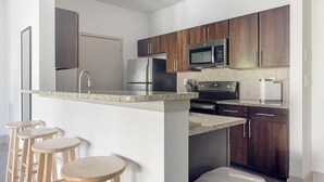 Una cocina moderna con armarios de madera oscura, electrodomésticos de acero inoxidable y encimera de granito. Hay tres taburetes de madera en la barra.