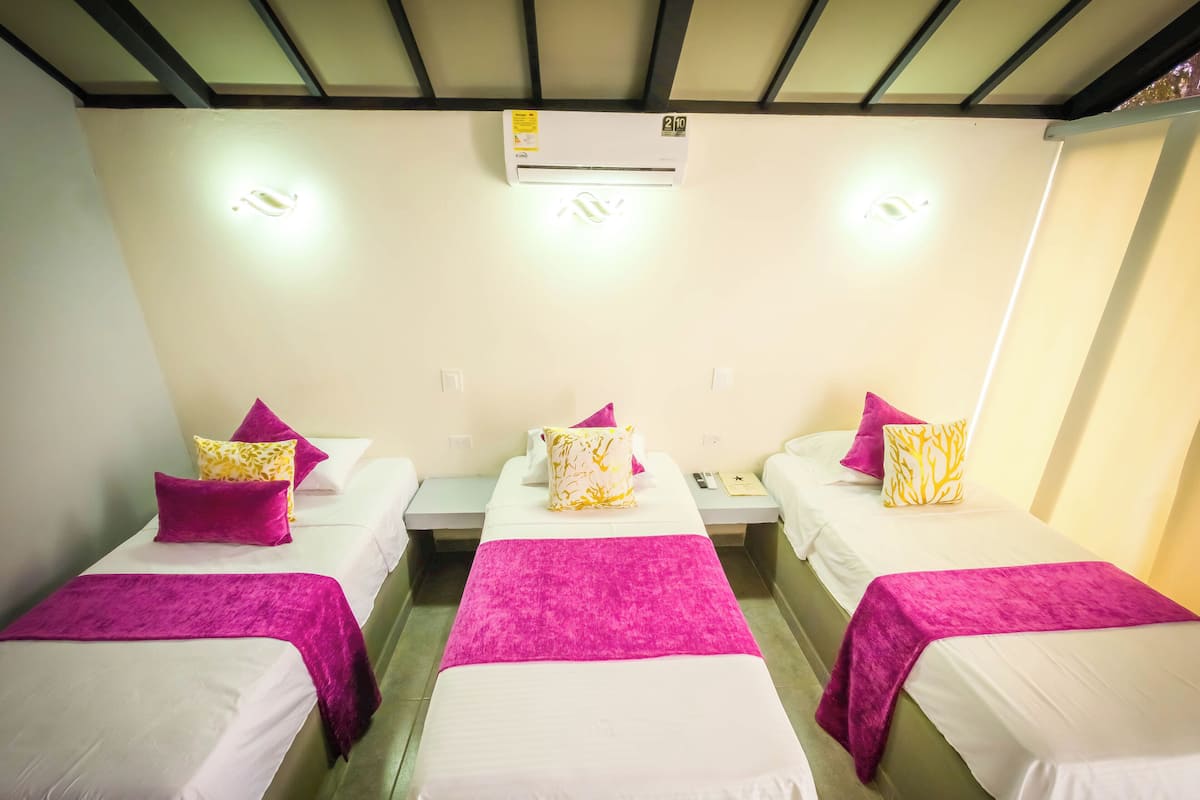 Habitación de hotel con tres camas individuales, cada una con colchón blanco y manta rosa. Hay un equipo de aire acondicionado sobre las camas, y una mesita entre ellas con un teléfono y un libro.