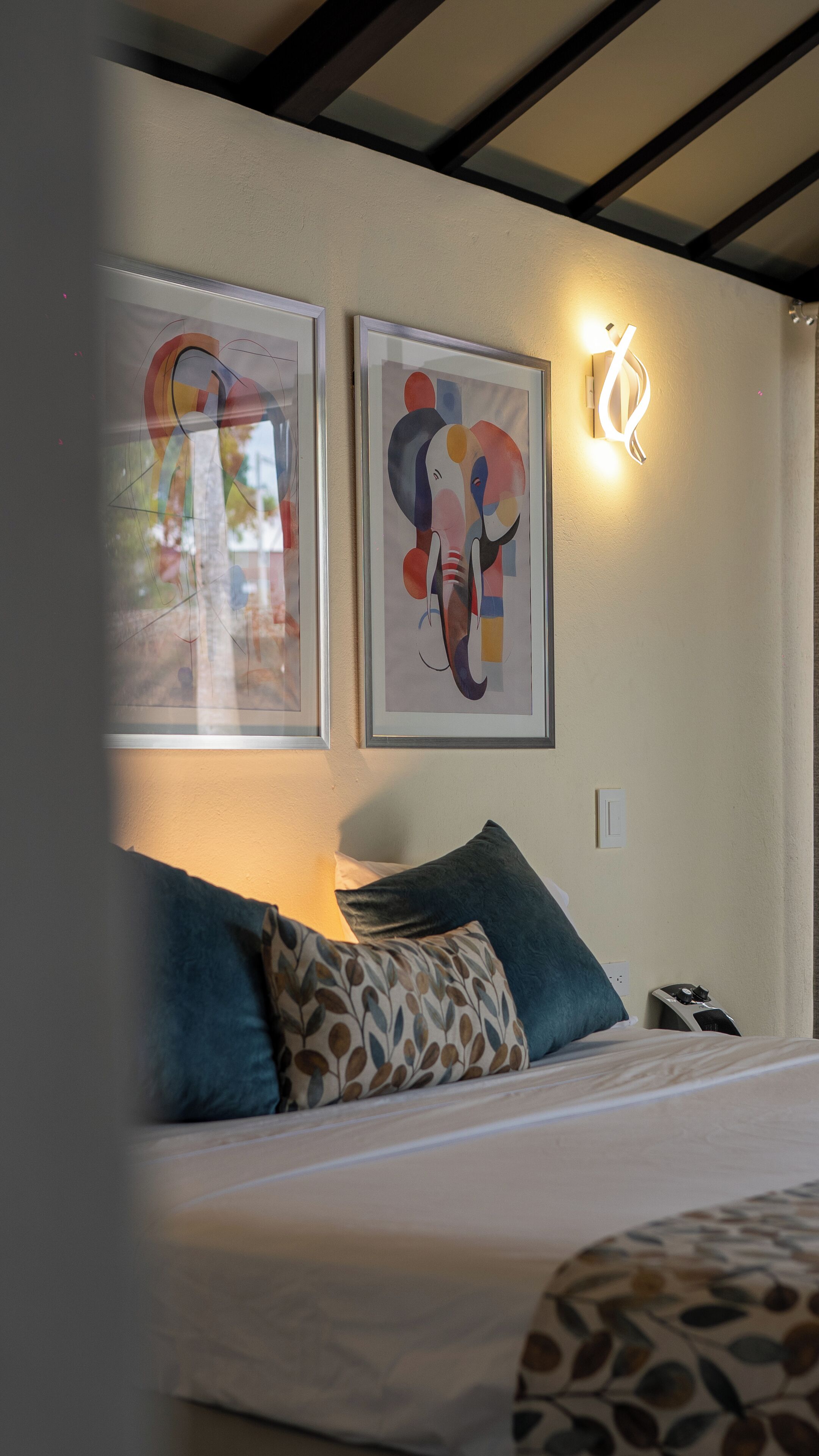 A hotel room with a bed, pillows, a wall-mounted light, and two framed abstract artworks.