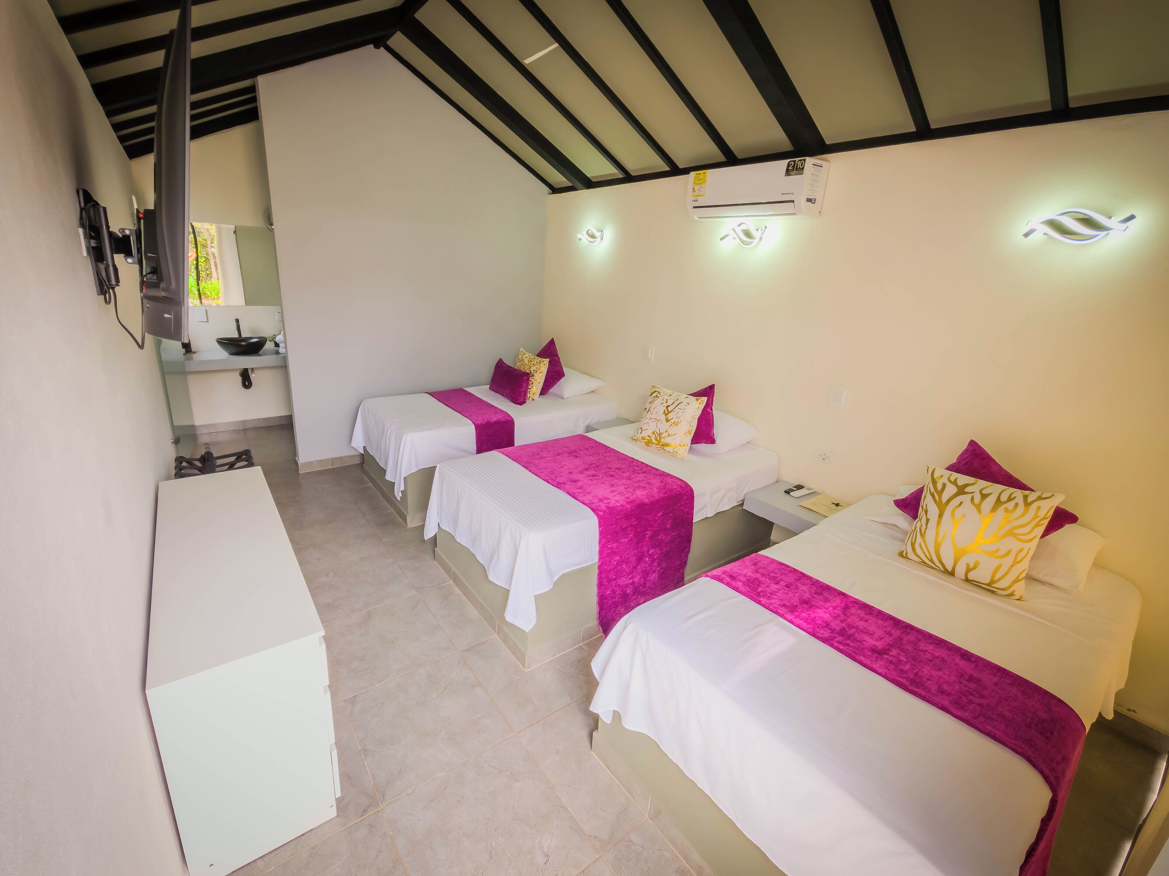 Ein Hotelzimmer mit drei Betten, jedes mit weißer Bettwäsche und rosa Akzenten. Es gibt einen kleinen Tisch und einen Stuhl, ein Waschbecken und einen Spiegel in der Ecke.