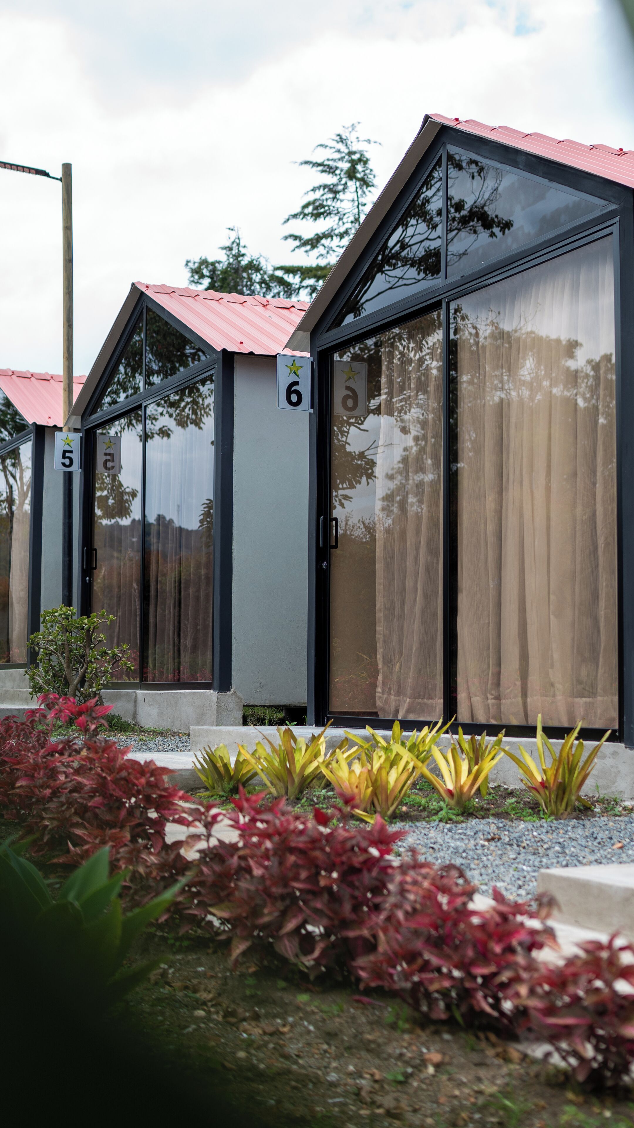 A row of modern, glass-fronted cabins with metal roofs, numbered 5, 6, and 7, surrounded by landscaped gardens.