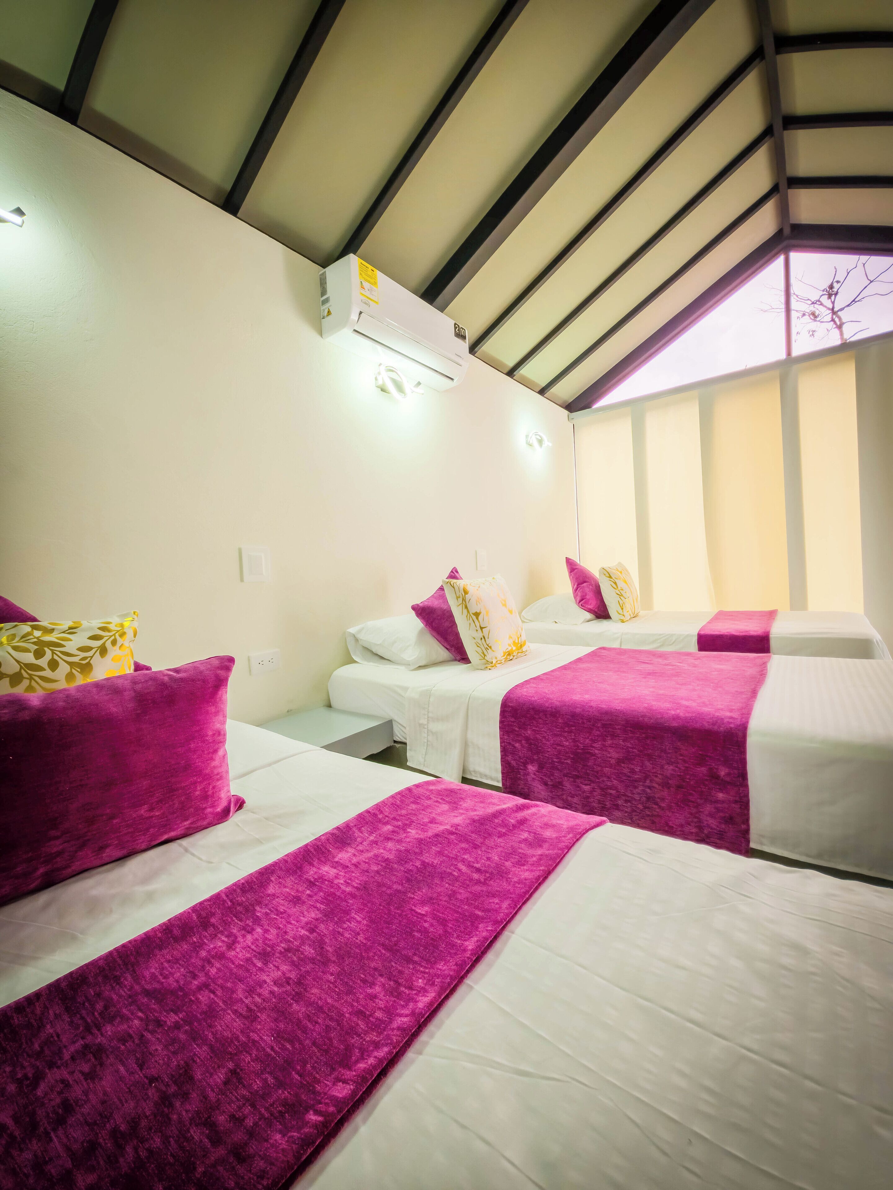 A hotel room with two beds, each with a white mattress and purple bedspreads. There is a ceiling fan and an air conditioning unit mounted on the wall.