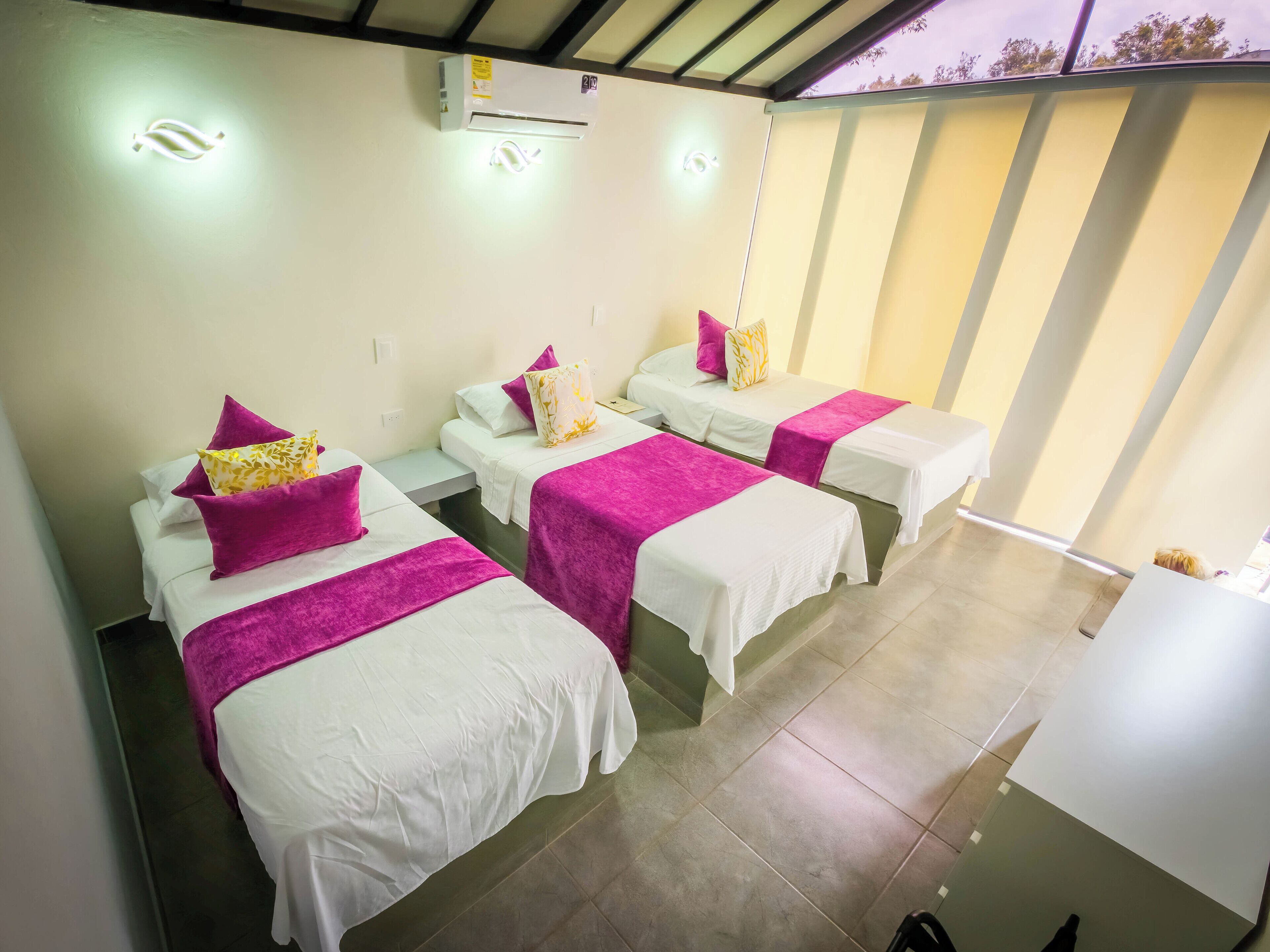 A hotel room with three beds, each with a white bedspread and a purple blanket. There is an air conditioning unit on the wall, and the room has a tiled floor.