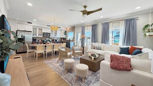 Ein modernes Wohnzimmer mit Sofa, Couchtisch und Poufs. Es gibt einen Essbereich mit Tisch und Stühlen. Im Hintergrund ist eine Küche mit weißen Schränken und einem Kühlschrank zu sehen.