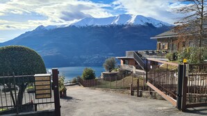 A gated entrance with a gravel path leading to a modern building with a stone facade and a metal gate. - CASA "INCANTO" Charming house with breathtaking views of Lake Como. (Comun)