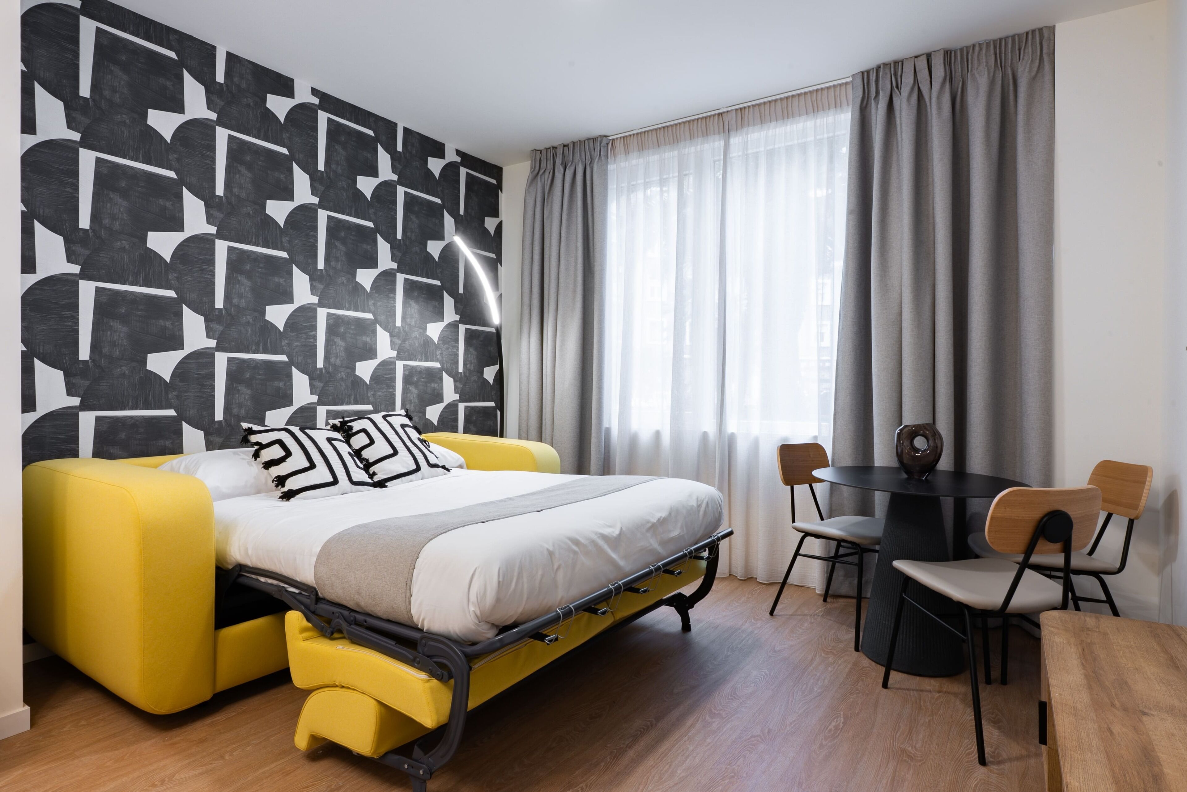 Una habitación moderna con sofá cama tipo futón, una mesa de comedor con sillas y un papel tapiz con motivos geométricos.