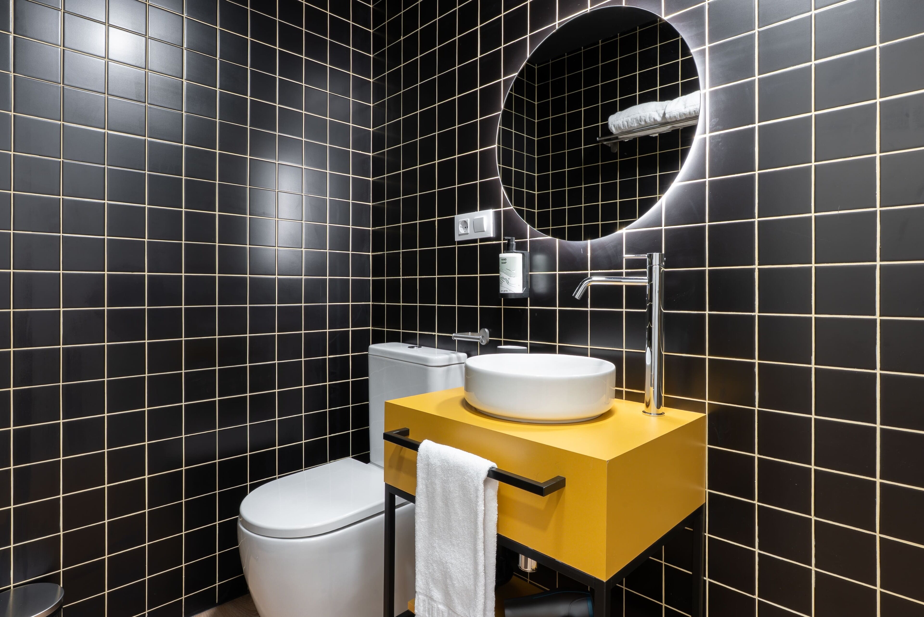 A modern bathroom with black tiled walls, a white toilet, a yellow vanity with a white sink, and a mirror above the sink.