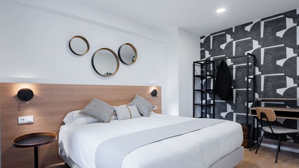 A modern hotel room with a large bed, a desk with a chair, and a coat rack. - Smartr Valencia Turia (Valencia)