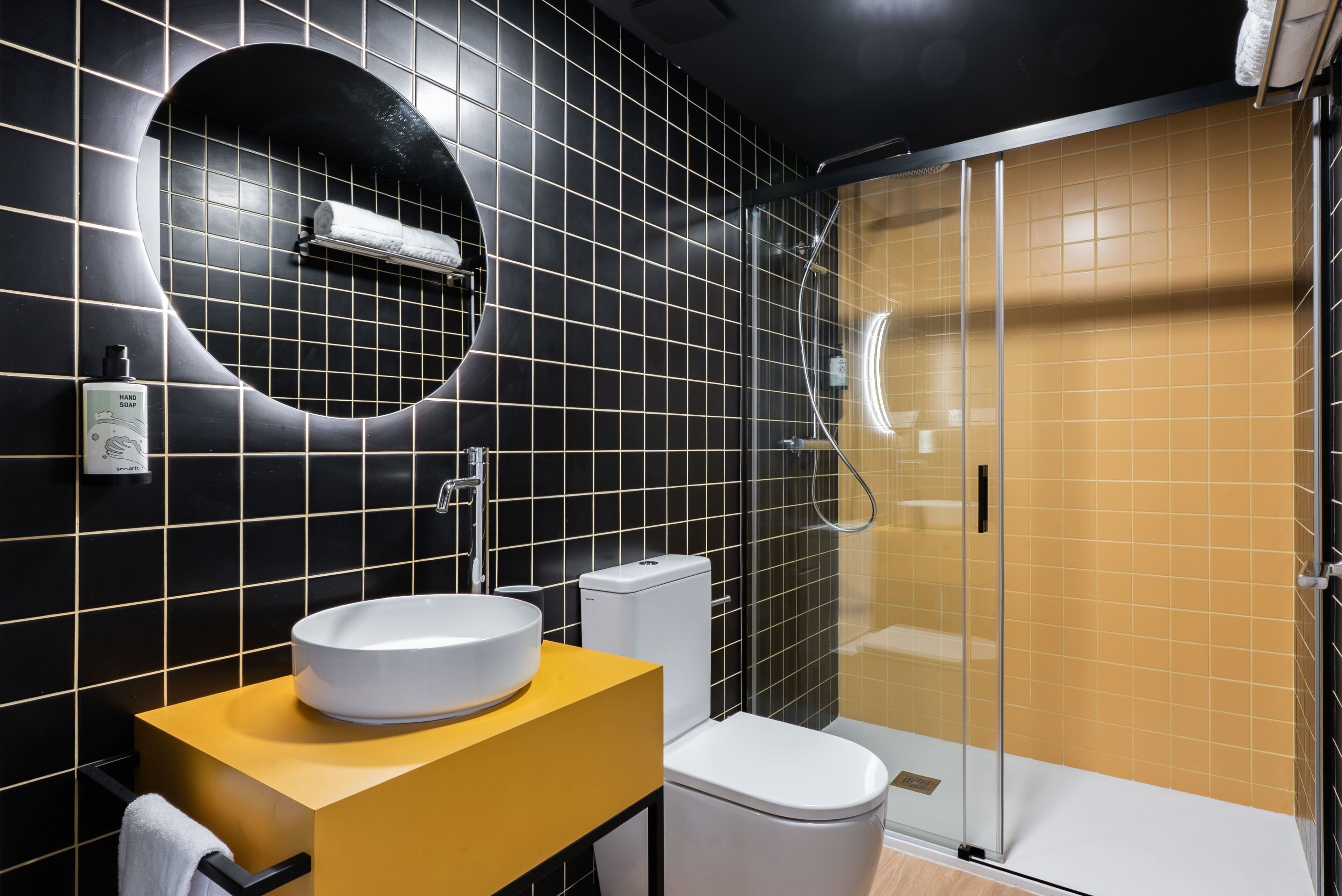 Un baño moderno con un revestimiento de azulejos negros y amarillos, un lavamanos blanco y una ducha con mampara de vidrio.