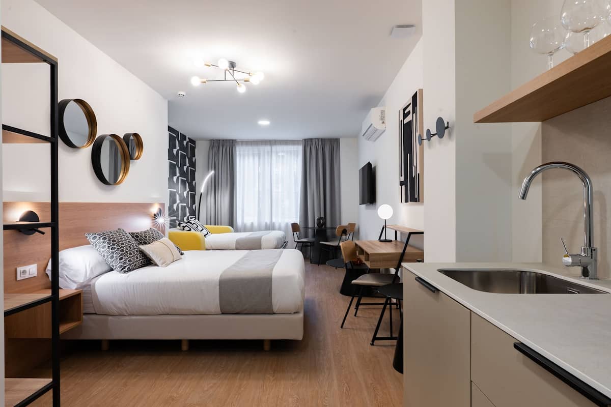 A modern hotel room with a bed, bedside tables, a desk, and a kitchen area with a sink.