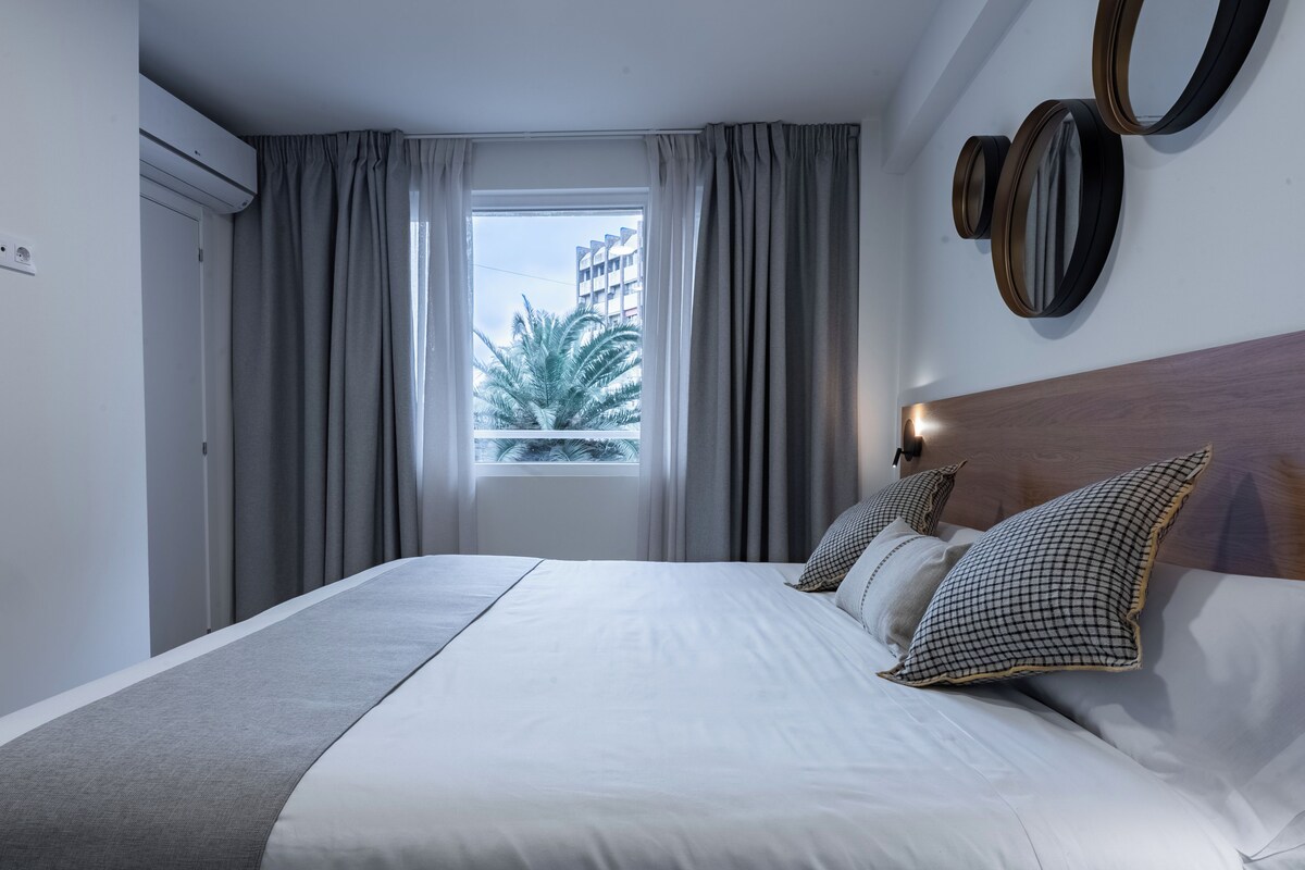 A modern hotel room with a large bed, grey curtains, a window with a view of palm trees, and a wooden headboard.