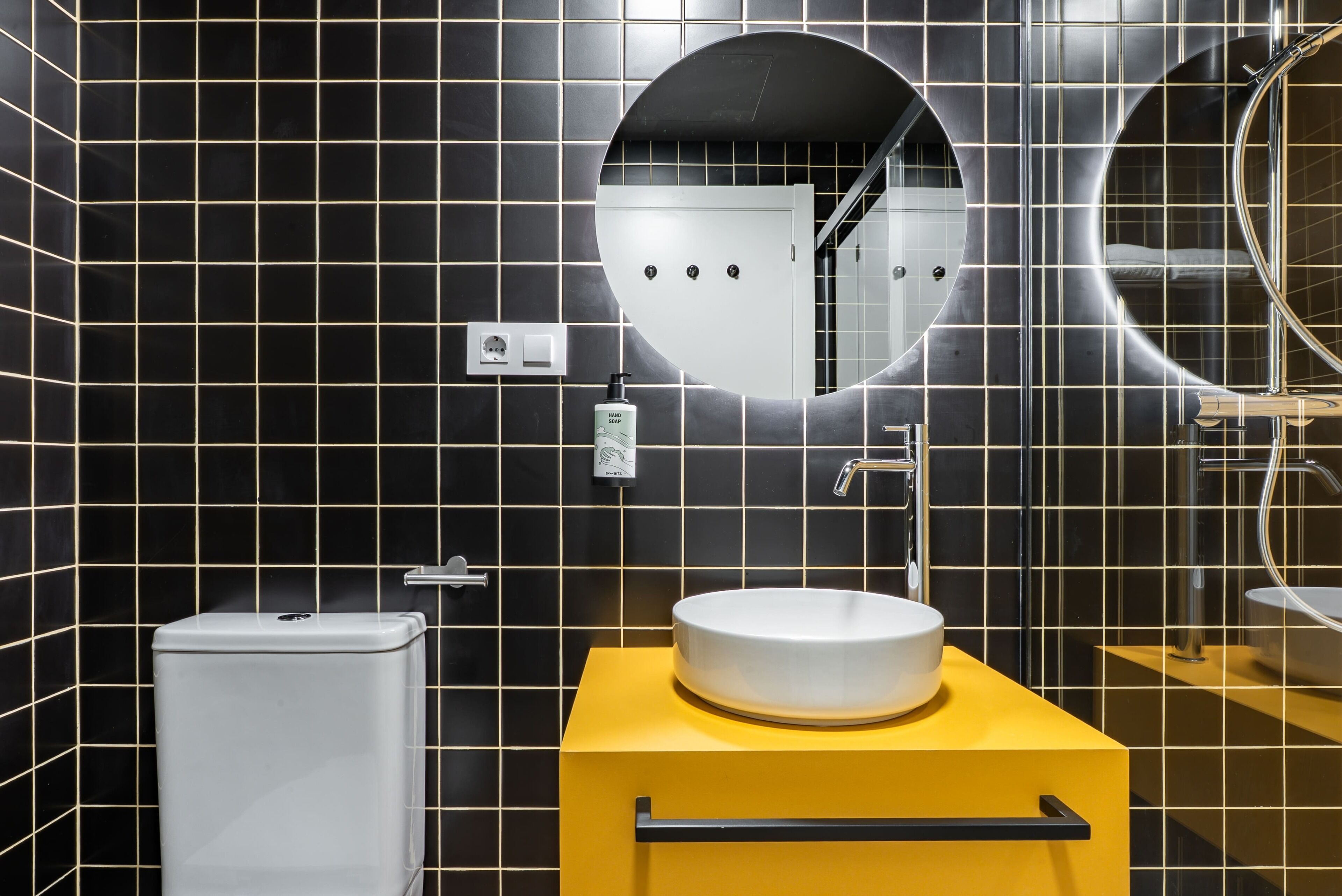 A modern bathroom with black tiled walls, a white toilet, a yellow sink vanity, and a large mirror.