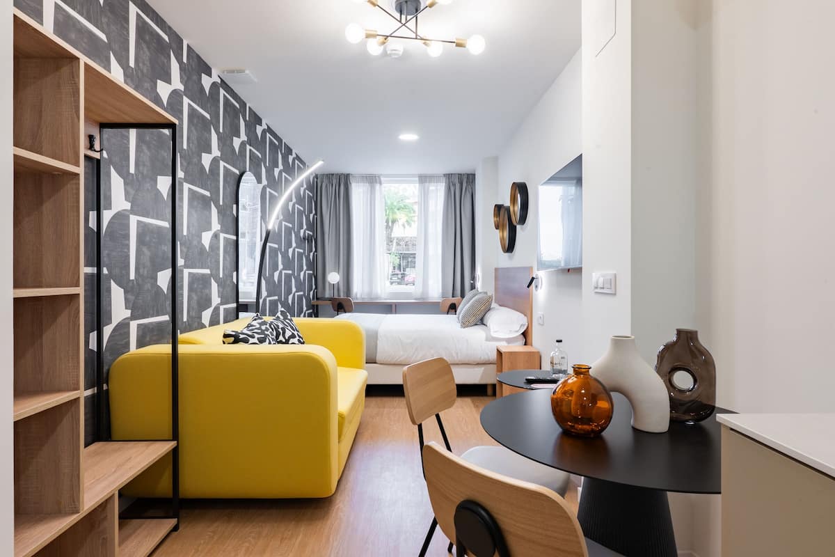 A modern hotel room with a yellow sofa, a bed with a white headboard, a dining table with decorative vases, and a geometric-patterned wall.
