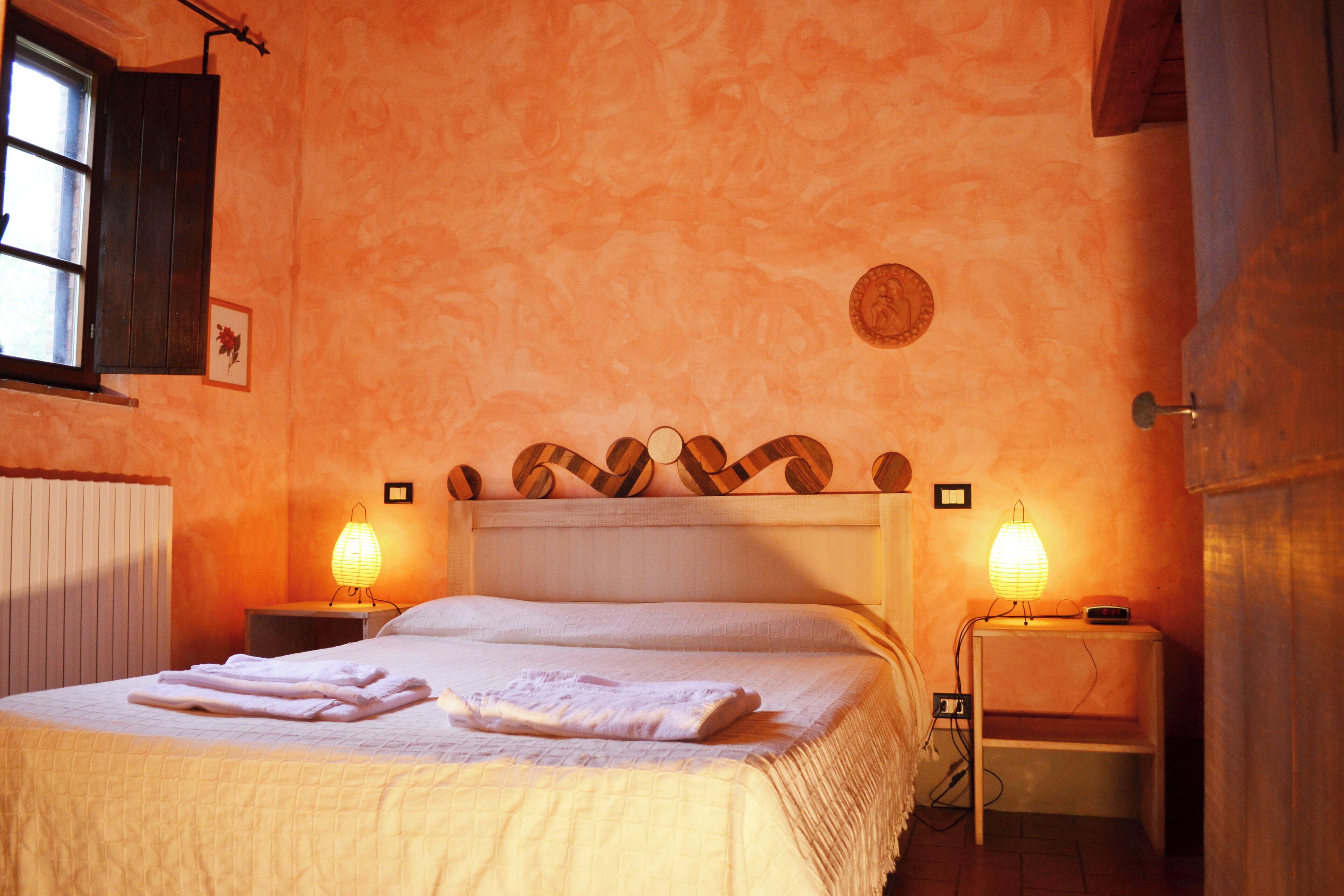 Ein ordentlich gemachtes Bett mit weißen Leinen und einem gefalteten Handtuch, zwei Nachttischlampen, ein Holzkopfschild und ein Fenster mit dunklen Jalousien.