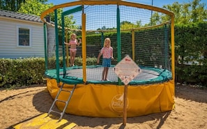 Un trampoline doté d’un filet de protection, entouré de sable, avec un panneau de signalisation jaune et un logo au sol.