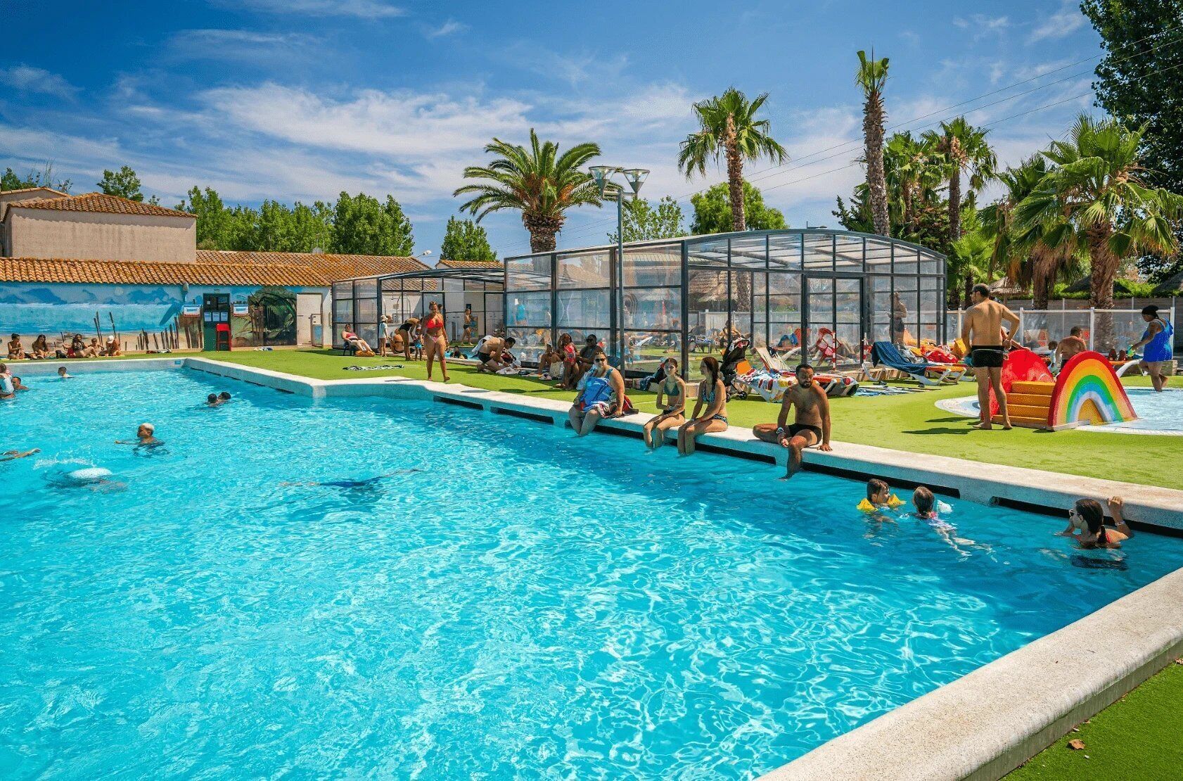 A swimming pool with people, palm trees, and a glass-enclosed structure.