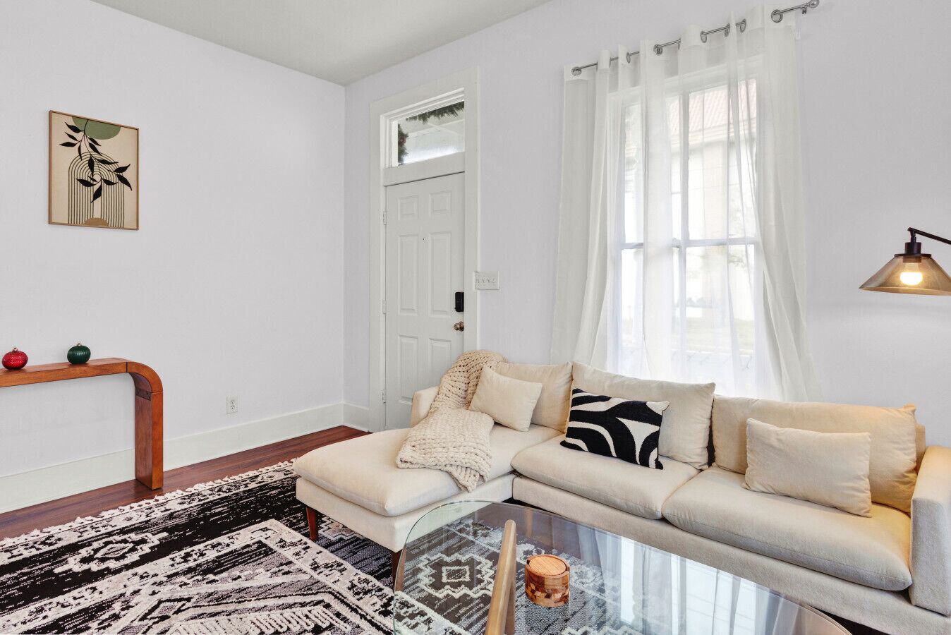 A living room with a sofa, a glass coffee table, a patterned rug, a wooden console table with decorative items, and a door with a window.