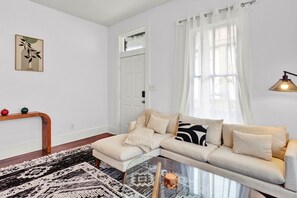 Un salon comprenant un canapé, une table basse en verre, un tapis à motifs, une console en bois agrémentée d’objets décoratifs et une porte avec une fenêtre.