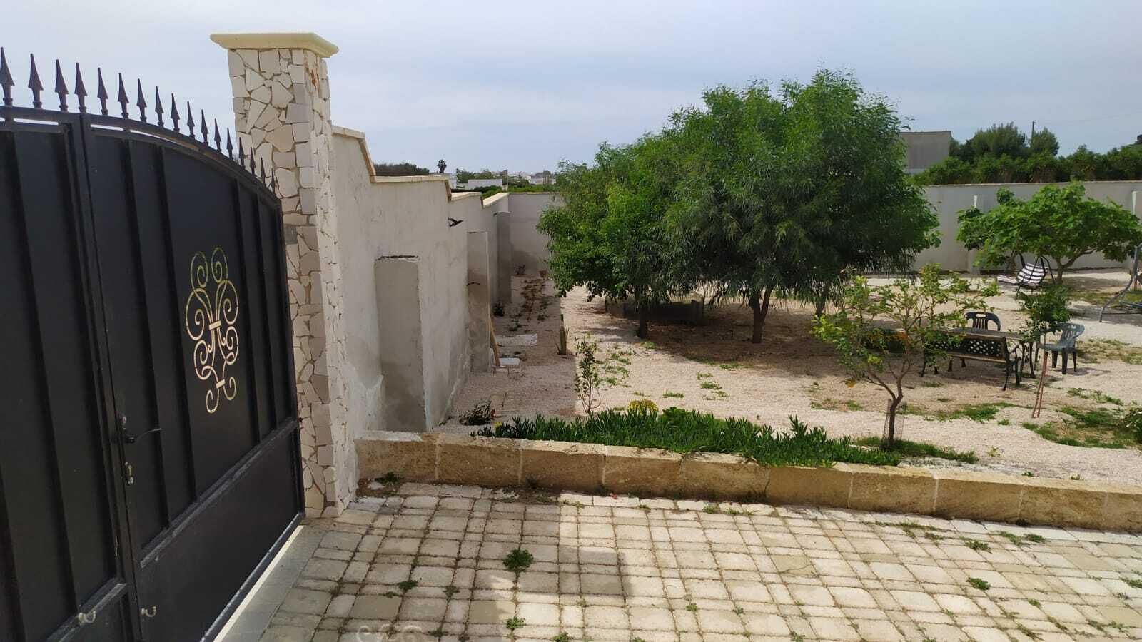 Une entrée fermée par un portail, avec un mur en pierre et un chemin pavé menant à un petit jardin agrémenté d’arbres et de bancs.