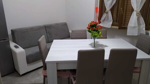Un pequeño comedor con una mesa blanca, cuatro sillas, un sofá gris y un florero con flores rojas.