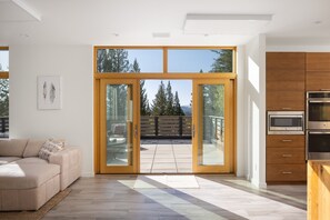 Une cuisine moderne avec des meubles en bois et des appareils électroménagers en acier inoxydable, une porte-fenêtre coulissante donnant sur un patio avec vue sur les arbres et les montagnes.