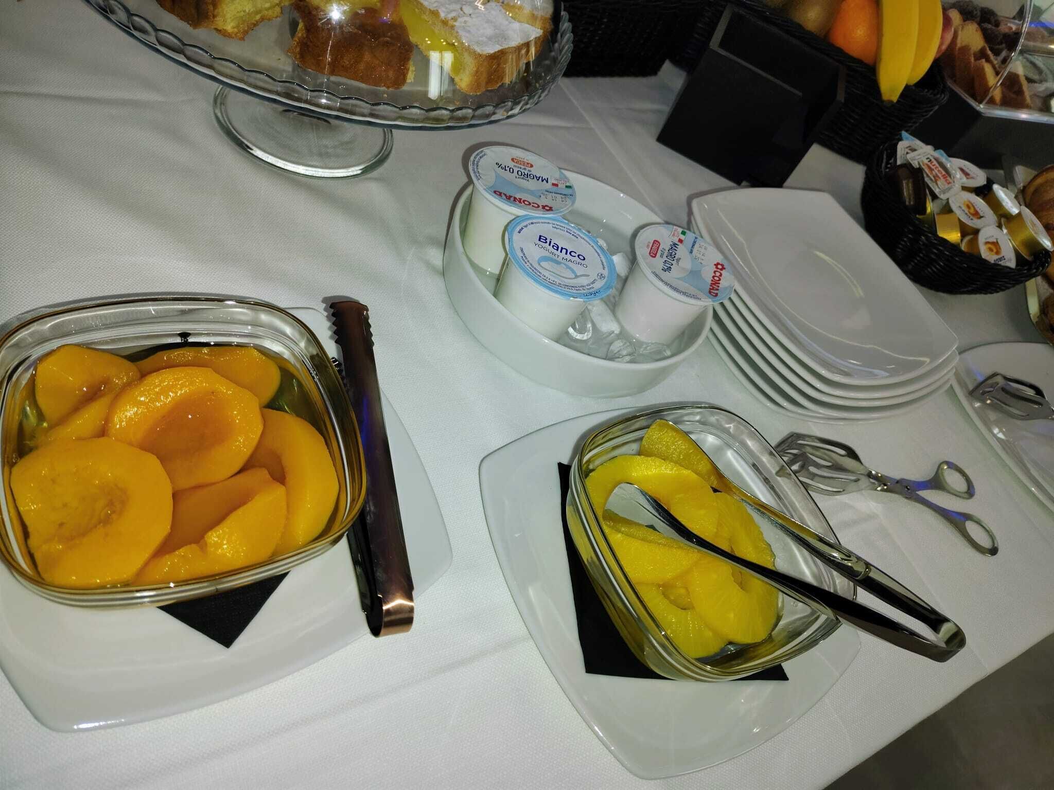 A buffet table with various food items including a dish of sliced peaches, bowls of yogurt, and a basket of bread.