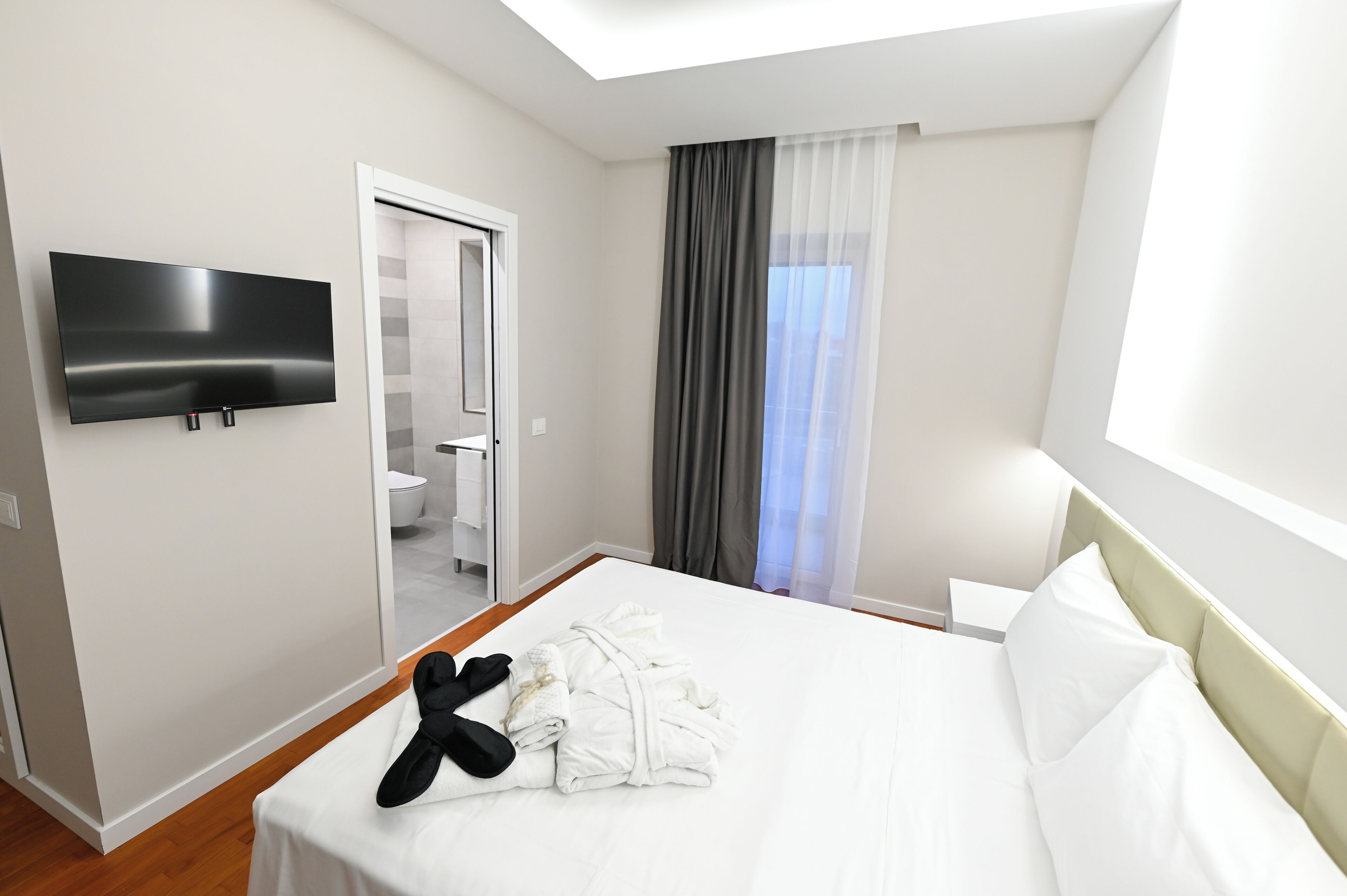 Quarto de hotel moderno com televisão de tela plana na parede, cama bem arrumada com lençóis brancos, um par de chinelos pretos e um banheiro visível através de uma porta aberta.