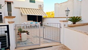 A gated patio area with outdoor seating, a tiled floor, and a white wall with a window and air conditioning unit.
